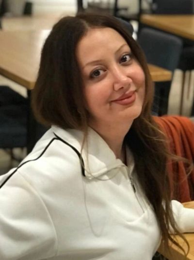 Smiling woman with long hair in a white jacket sitting indoors.