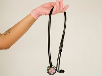 Hand wearing pink glove holds a black stethoscope against a plain background.