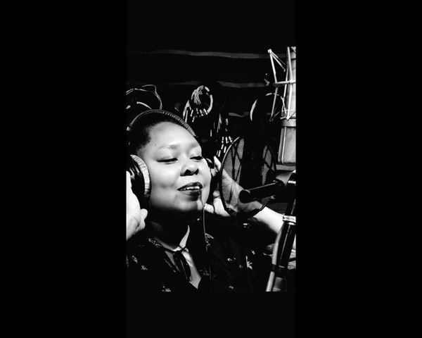 Sheryl Denise in a recording booth pressing headphones to her ears and beginning to speak in the mic