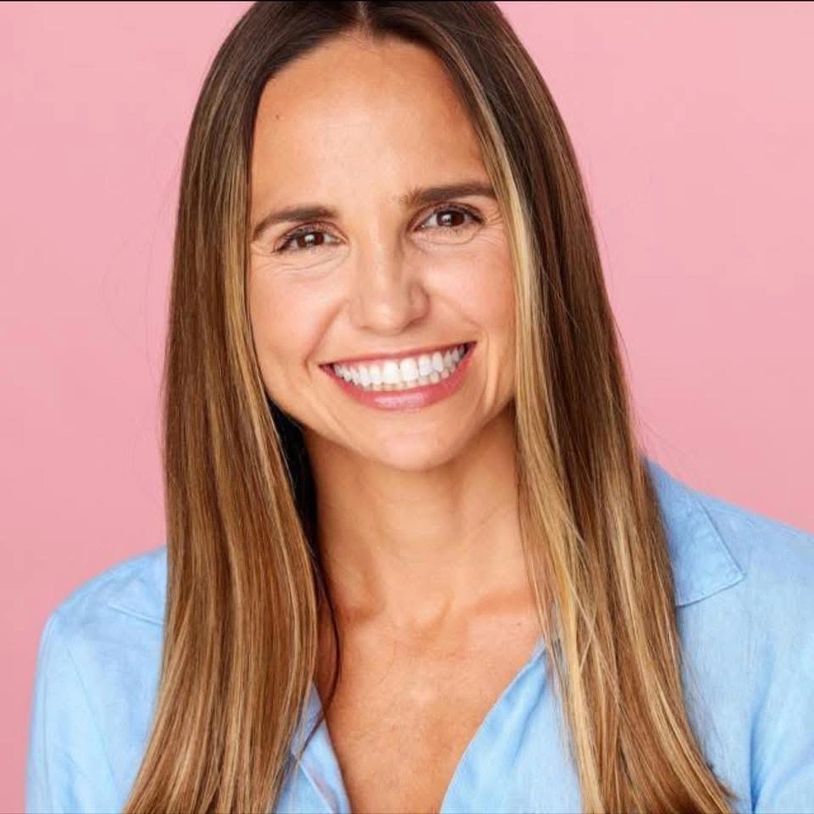 Smiling woman with long brown hair and blue shirt against pink background.