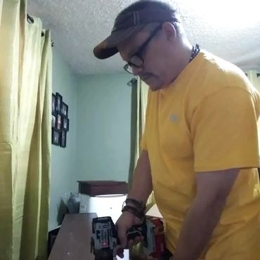 Man in yellow shirt using a power drill indoors on a wooden surface.