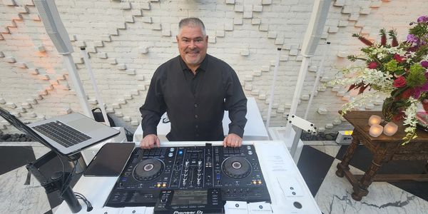 A smiling DJ stands behind a mixer setup with a laptop and flowers nearby.