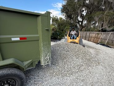 Man operating a small excavator spreading gravel from a trailer.
