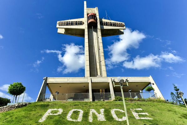 Large concrete cross monument with 'PONCE' spelled in stones on grass under a bright blue sky.