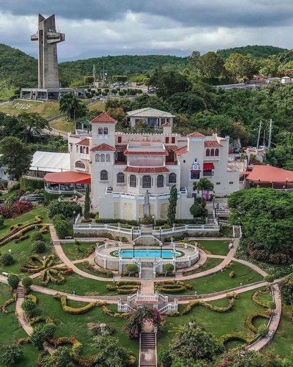 A grand white mansion with red roofs surrounded by manicured gardens and a large monument in the background.