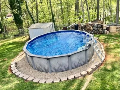 Above-ground pool with clear blue water in a backyard surrounded by trees.