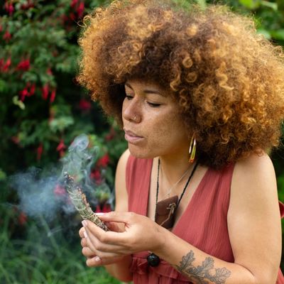Picture of a healer of mixed heritage blowing on a handmade smudge stick of mugwort, sage and rose.