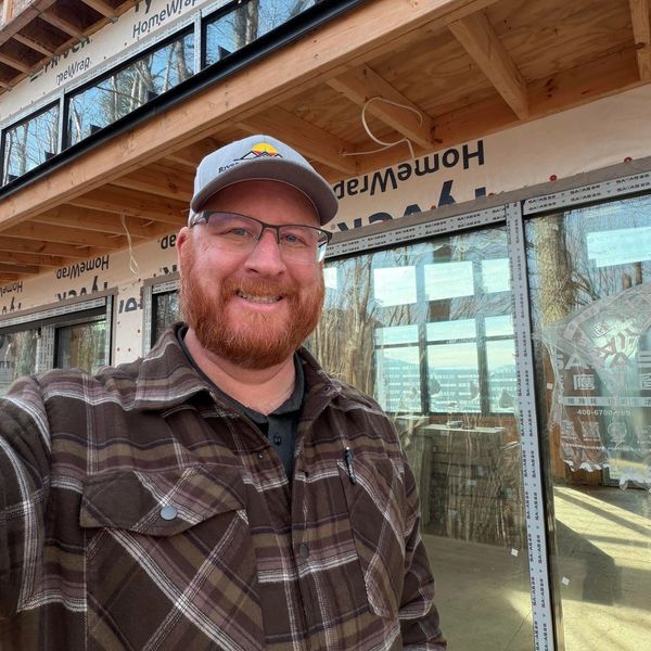 Smiling man in plaid shirt and cap at a wooden construction site.