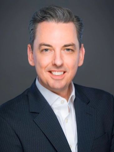 Professional man in a dark suit smiling against a gray background.