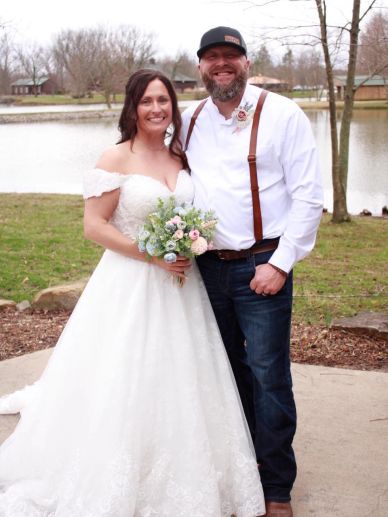 Bride in a white dress and groom in casual wedding attire pose happily outdoors.