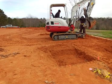 Two workers operate a compact excavator on a red dirt construction site.