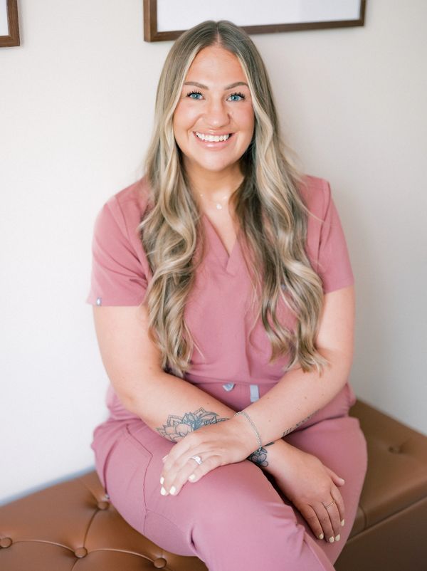 Smiling woman with long blonde hair in mauve scrubs sitting on a bench.