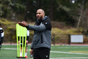 A coach gestures while holding a stopwatch on a sports field.