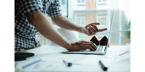 Person using laptop with small house models on desk near window.