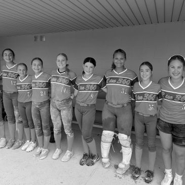 A girls' softball team lined up in uniforms, smiling in a dugout.