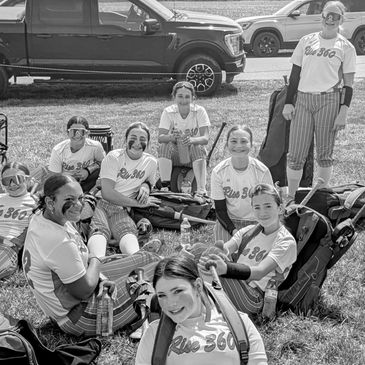 A group of female softball players resting on grass, wearing matching uniforms and masks.