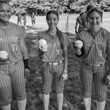Three young softball players smile, holding baseballs and wearing matching jerseys.