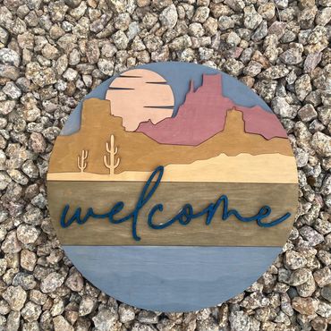 Wooden welcome sign with desert landscape and cacti on gravel.