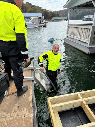 Two men in bright yellow jackets by the water, one climbing out of the lake using a ladder.