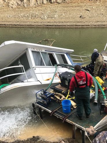Workers pumping water from a partially submerged houseboat to prevent sinking.