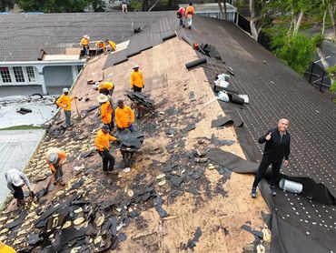 Workers in orange shirts remove old roofing materials from a large roof.