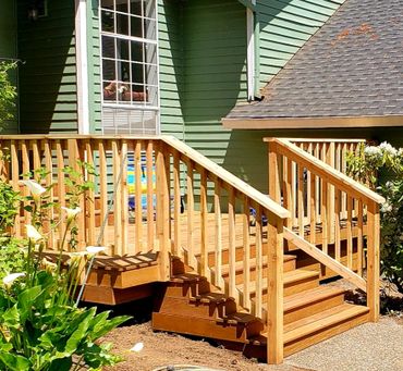 Newly built wooden deck with stairs and railings outside a green house.