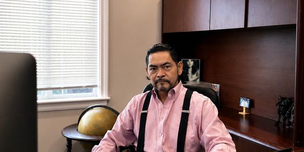 Hombre serio sentado en oficina con camisa rosa y tirantes negros.