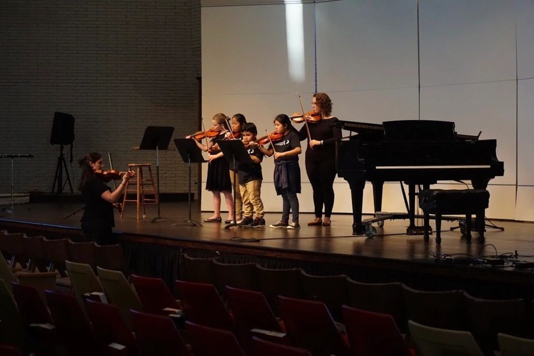 Xaris Emmett leading a group class of beginner violin students on stage.