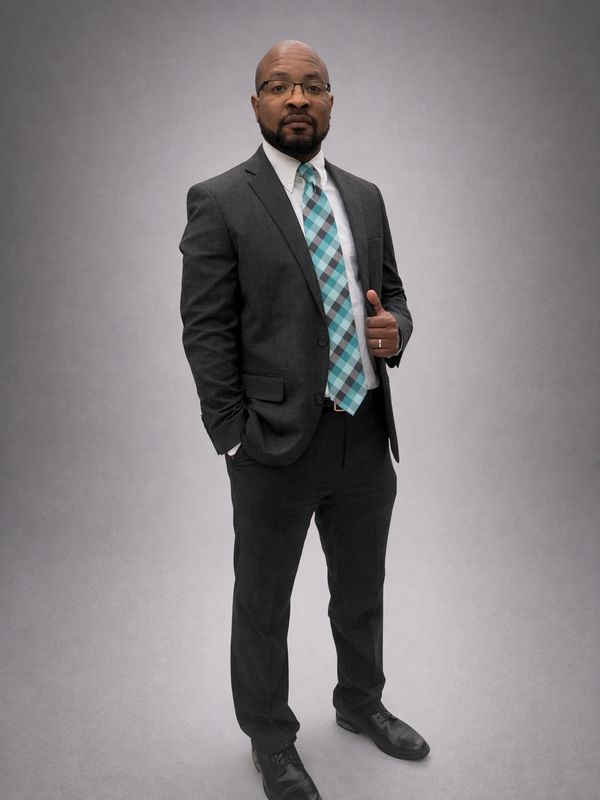 Man in a dark suit with a blue plaid tie standing confidently.