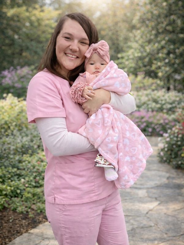 Woman in pink scrubs holding a baby wrapped in a pink blanket outdoors.