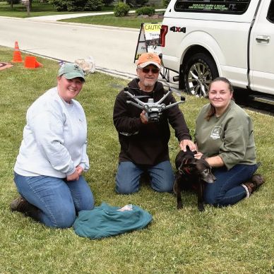 Three people kneeling on grass with a dog and holding a drone.