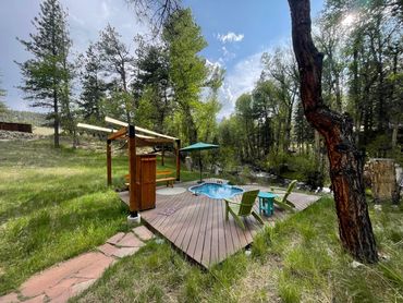 Blue skies, sunlit trees, green grass, & view of hot spring on decking next to Chalk Creek