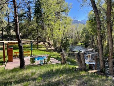 Sunlit trees & view of hot spring on decking with shade, seating and hammock along Chalk Creek