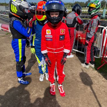 Young go-kart racers in colorful racing suits and helmets prepare for a race on a sunny day.