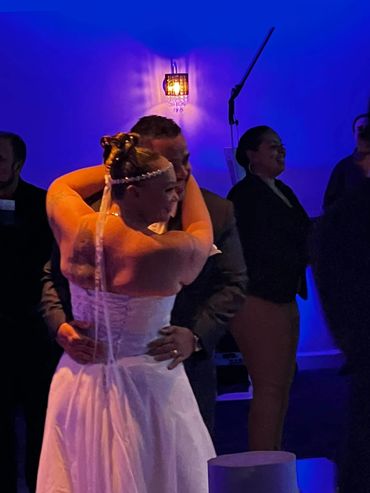 A couple shares an emotional dance at a dimly lit event with blue ambient lighting.
