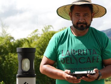 Man in a green shirt and straw hat holding a tray, standing behind a table with coffee-related items.
