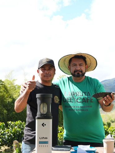 Two men showcasing coffee equipment outdoors, one giving thumbs up.