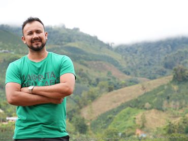 Smiling man in a green shirt stands with arms crossed against a scenic mountain backdrop.