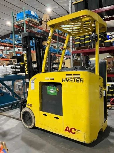 Yellow Hyster stand-on forklift in a warehouse with shelves and boxes.
