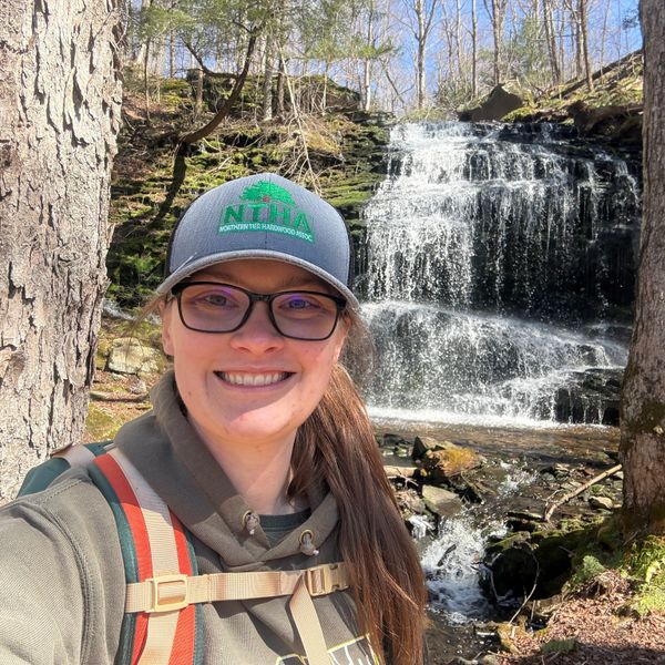 Smiling woman with glasses and hat hiking near a scenic waterfall in a forest.