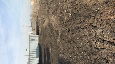 A barren dirt lot with tire tracks between fences and industrial structures.