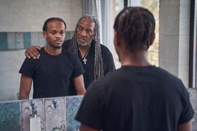 A young man looks into a bathroom mirror while an older man with long gray locs stands behind him, r