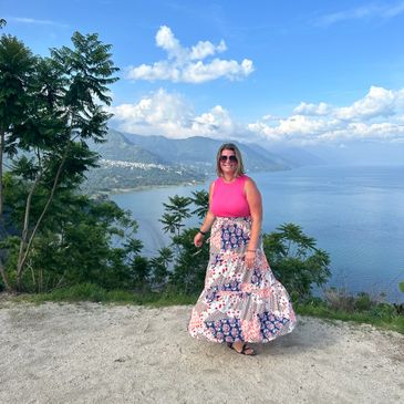 Woman in a vibrant skirt stands by a scenic lakeside viewpoint with mountains in the background.