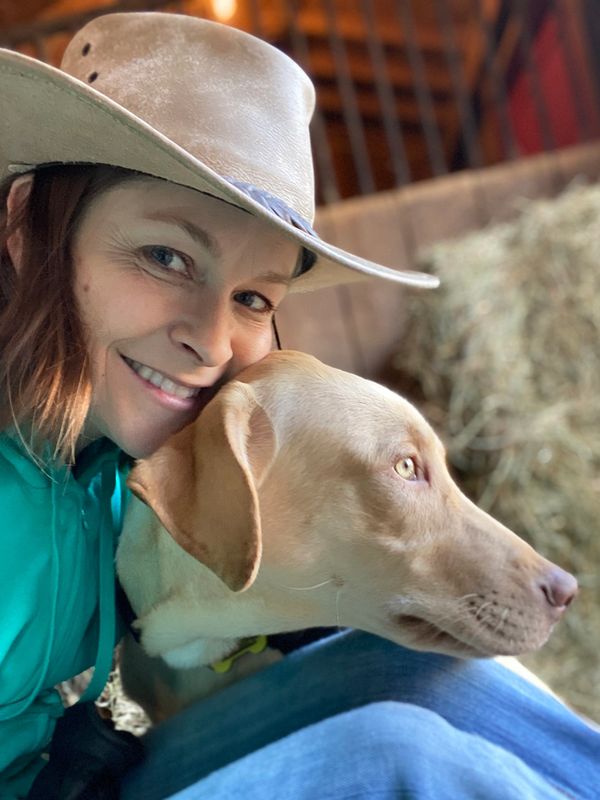 Woman in a cowboy hat smiling with her dog in a rustic barn setting.