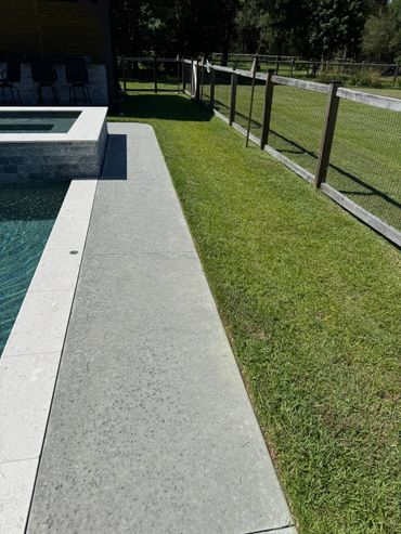 Concrete poolside walkway beside a grassy lawn and wooden fence.
