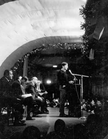 A black and white photo of a student speaking at a microphone before a group