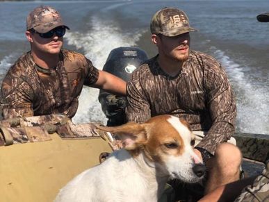 Two men in camouflage on a boat with a dog on the water.