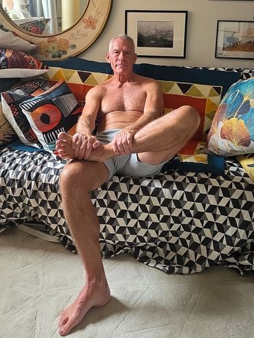 Shirtless man sitting cross-legged on a patterned couch with colorful pillows.