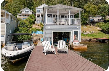 Two white chairs on a dock by a boathouse and a boat on a calm lake.