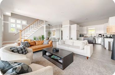 Bright, modern living room with beige and white furniture, wooden staircase, and open kitchen.